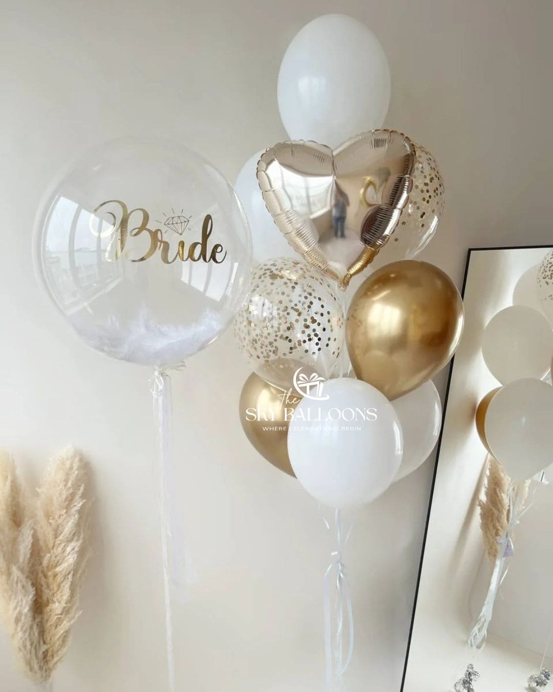 Bridal balloon arrangement with a clear “Bride” bubble balloon, a metallic gold heart, white and gold latex balloons, and clear gold confetti balloons.