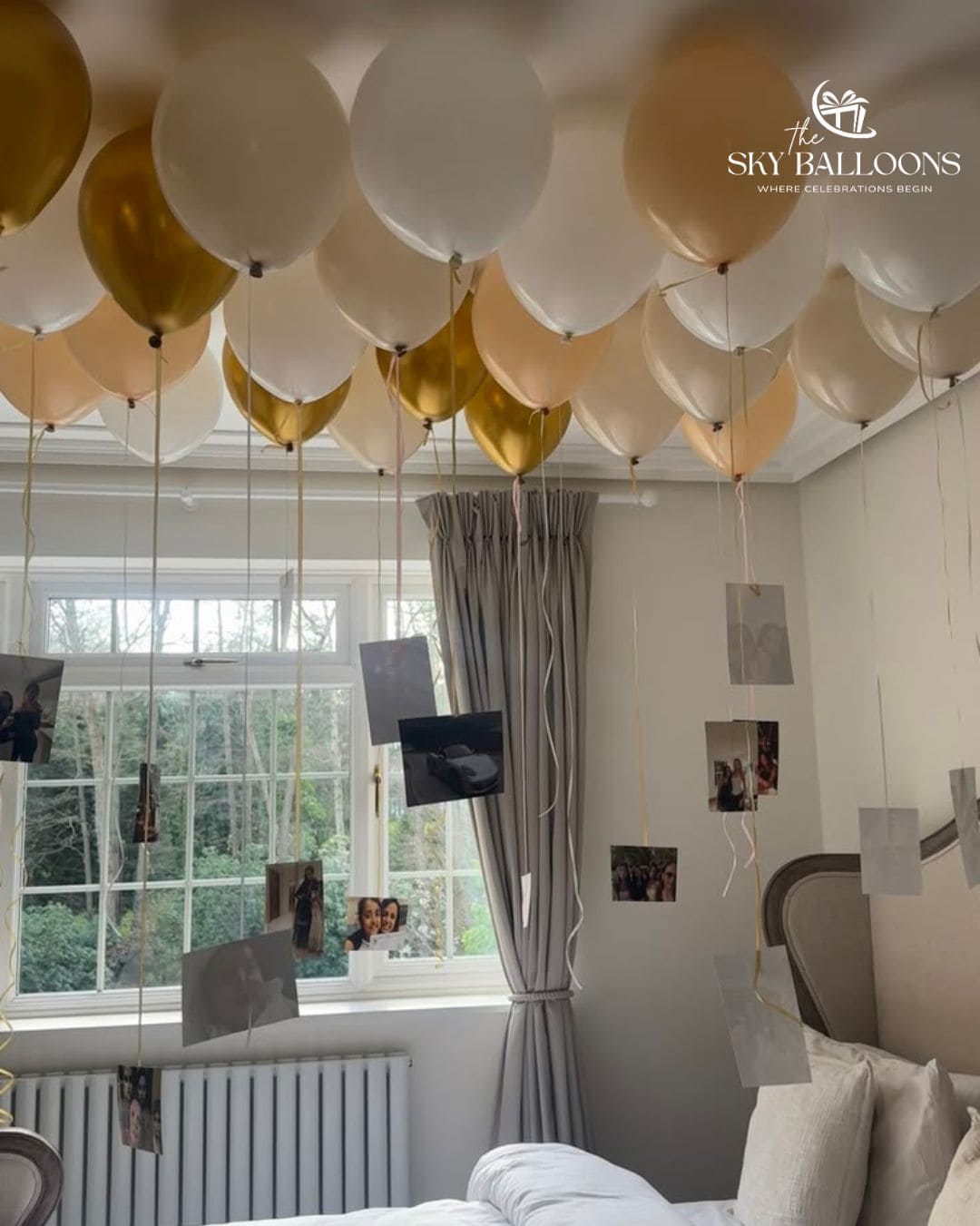 A ceiling balloon garland decoration featuring white, champagne, and metallic gold balloons suspended in a bedroom. The room has gray curtains, a bed with white bedding, and several photo prints hanging from strings on the wall.