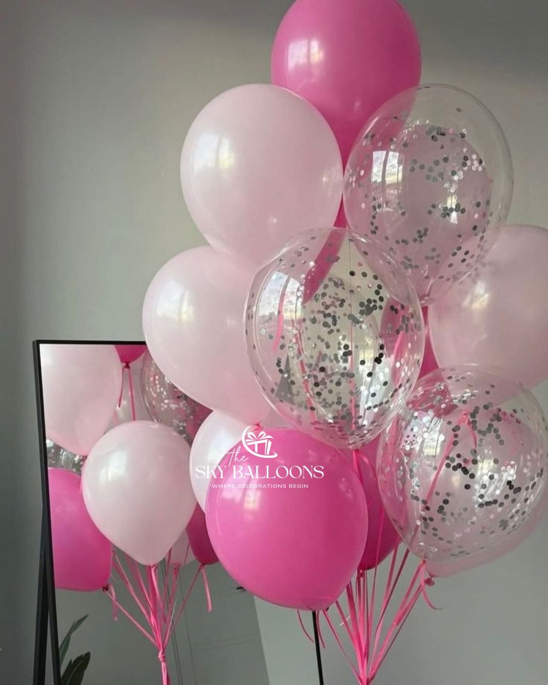 A cluster of pink latex balloons, including solid hot pink, pale pink, and clear balloons filled with silver confetti, tied together with pink ribbons. A mirror on the left reflects the balloons, and a 'The Sky Balloons' logo watermark is visible on one of the pink balloons.