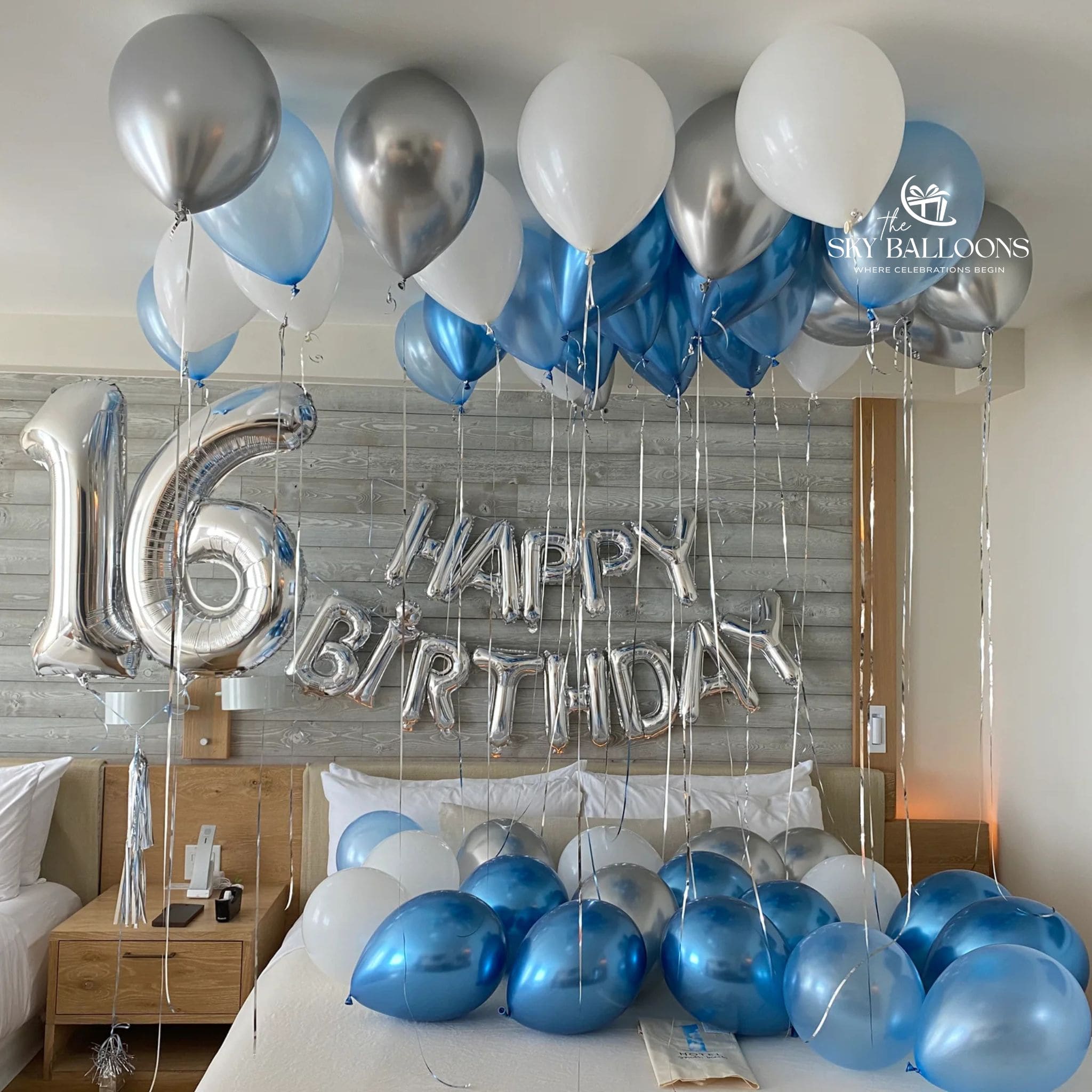 16th birthday bedroom decoration with silver number 16 and “Happy Birthday” foil balloons, plus blue, white and metallic silver balloons on the ceiling and bed.