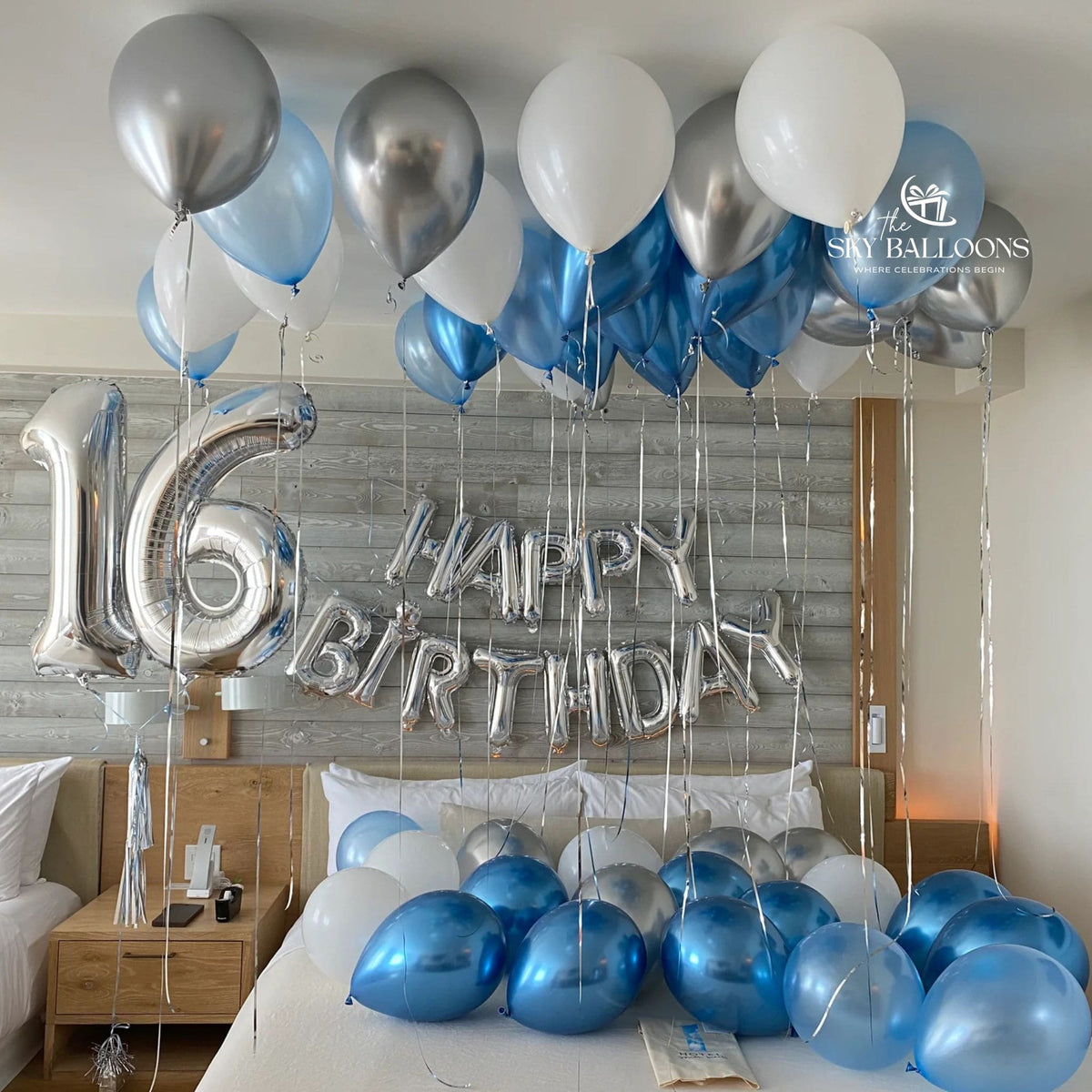 16th birthday bedroom decoration with silver number 16 and “Happy Birthday” foil balloons, plus blue, white and metallic silver balloons on the ceiling and bed.