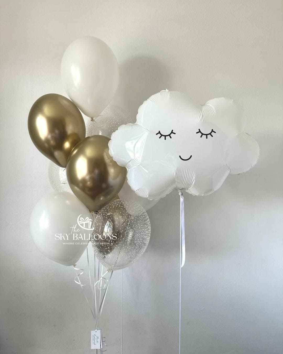 Smiling white cloud foil balloon beside a bouquet of white, metallic gold and clear confetti balloons with trailing white ribbons.