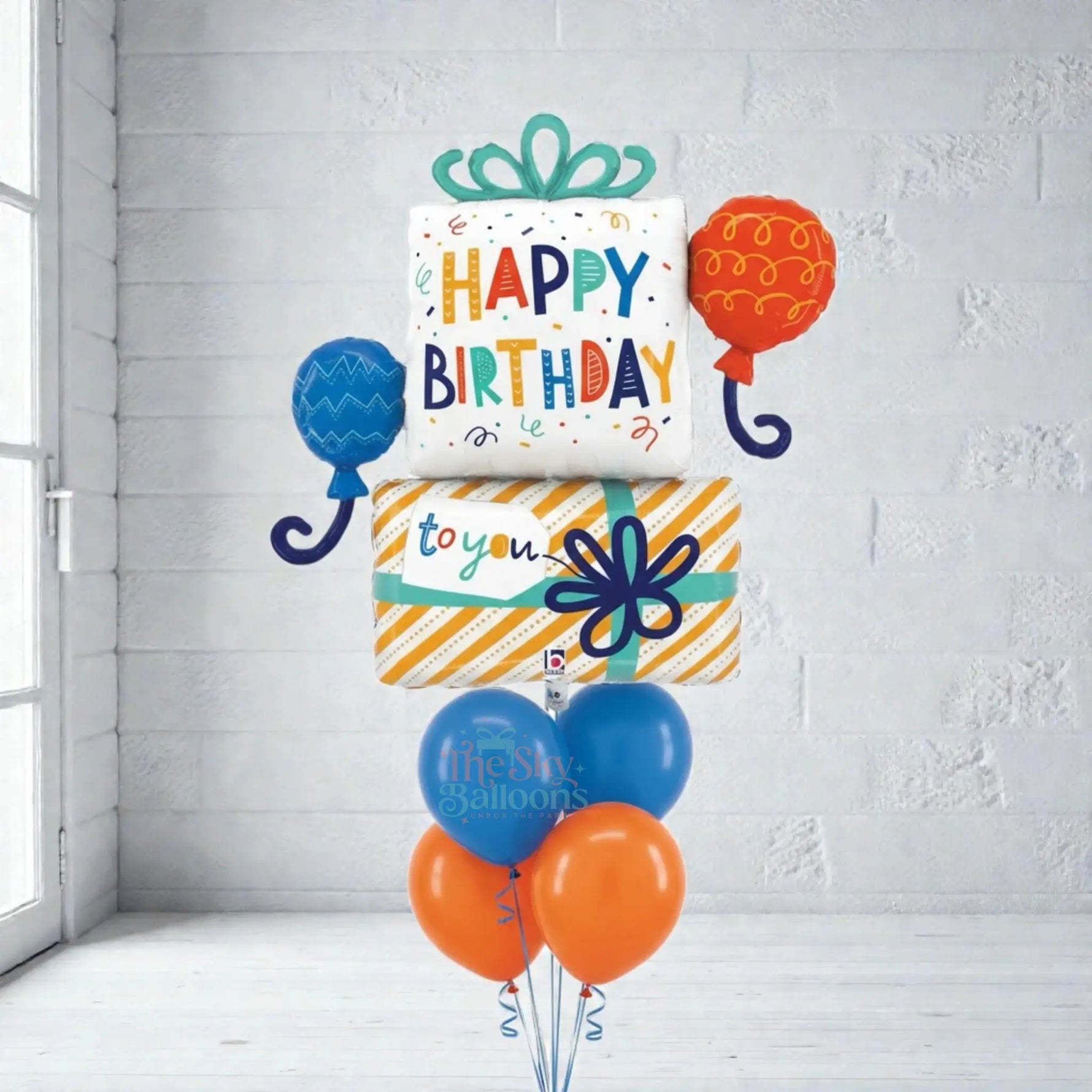 Colorful birthday balloon set with 'Happy Birthday' and 'To You' text against a white brick wall.