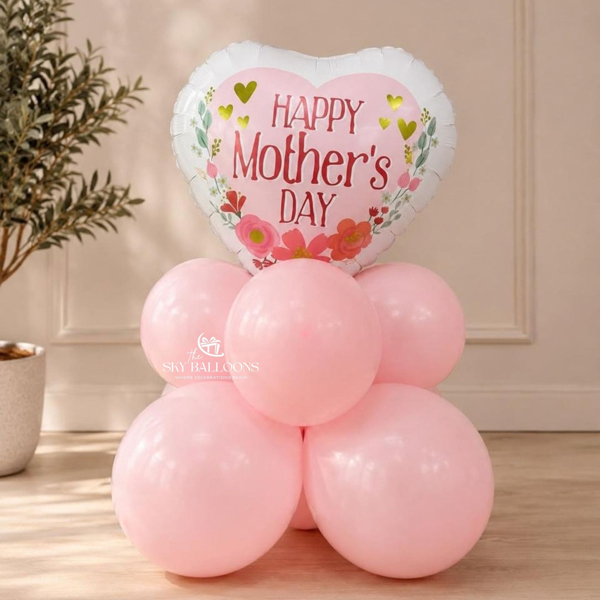 A pink heart-shaped foil balloon with 'Happy Mother's Day' text and floral graphics, floating above a cluster of six light pink latex balloons on a wooden floor in an indoor setting.