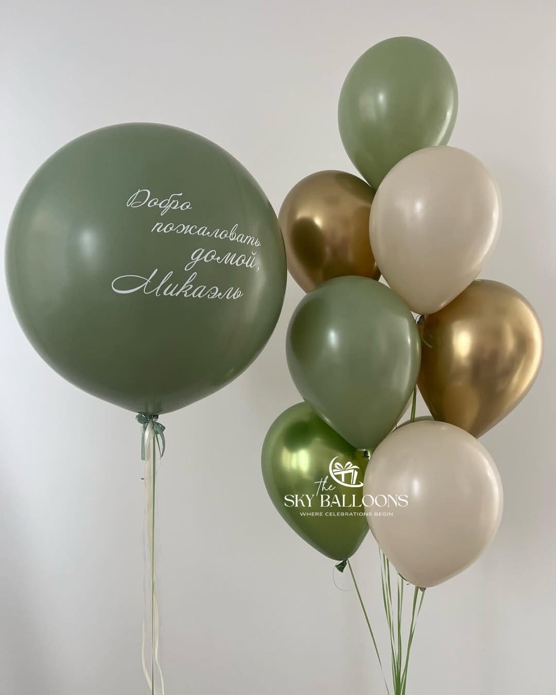 Personalised jumbo sage green balloon with a Russian welcome-home message beside a bouquet of sage green, cream and metallic gold balloons.