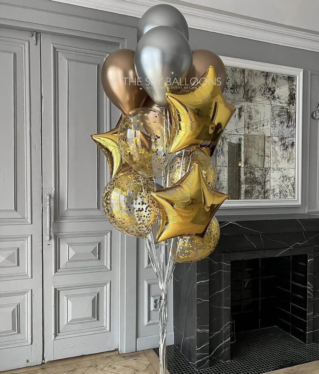 Decorative arrangement of gold, silver, and clear balloons in a room with a fireplace and framed picture on the wall.