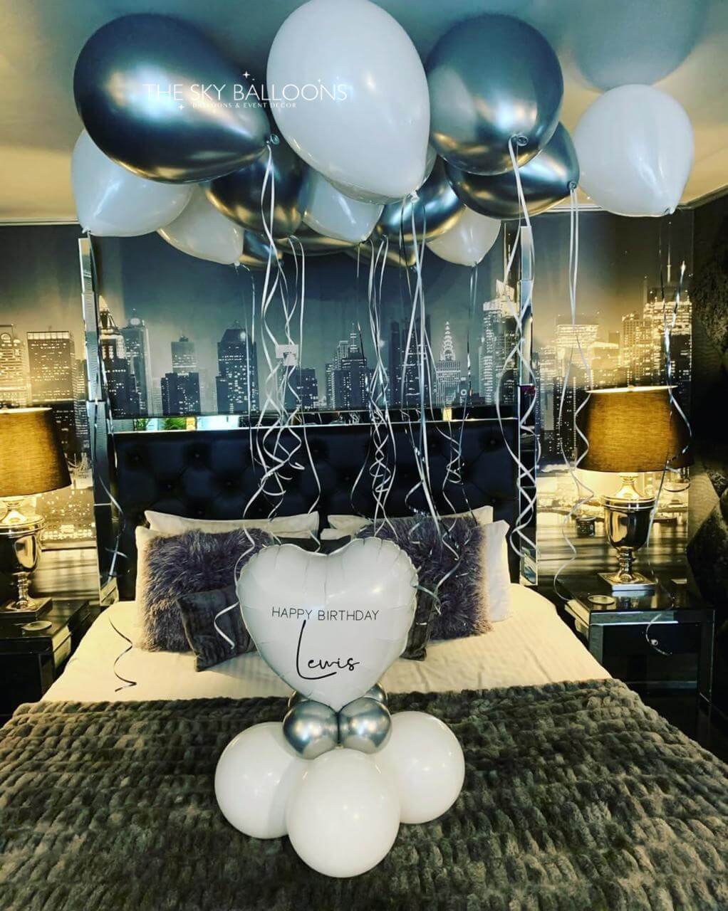 Decorative setup with balloons and a heart-shaped balloon on a bed in a room with cityscape wallpaper.