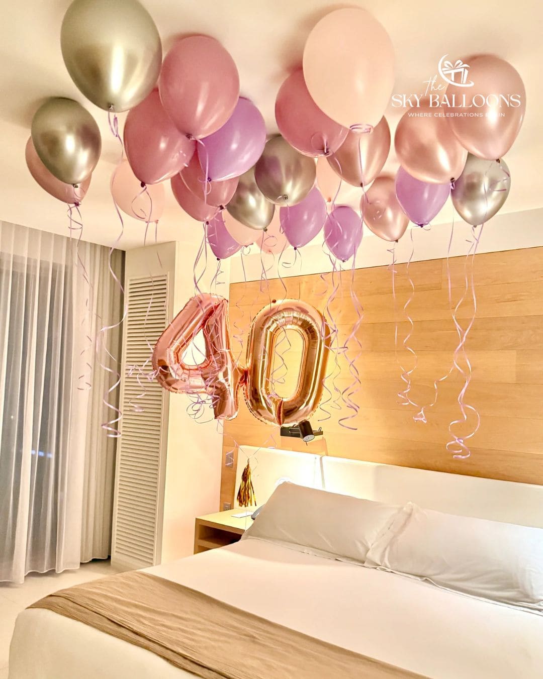 A bedroom decorated with a cluster of pastel pink, lavender, and metallic rose gold balloons, including two large foil balloons spelling out '40'. The room features a light wood headboard, white bedding, and sheer curtains.