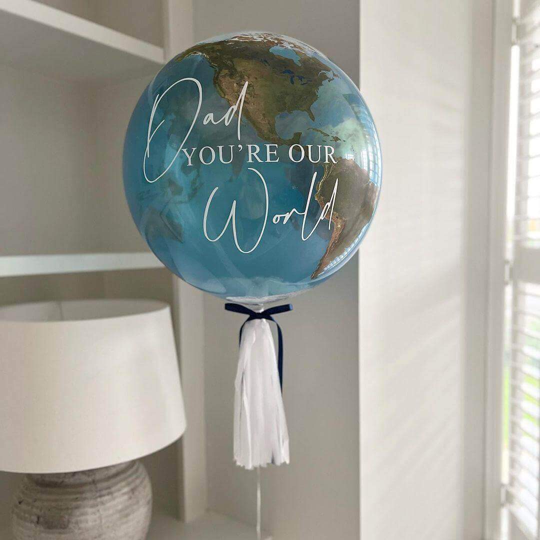 A balloon with a design resembling the Earth's globe and the text 'Dad you're our world' printed on it, adorned with tassels.