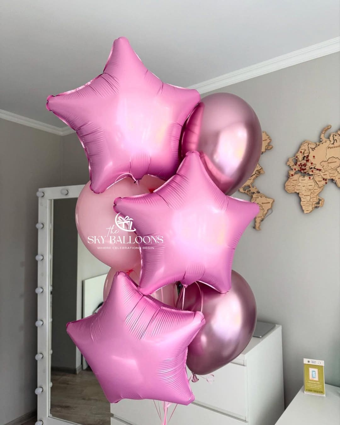 A cluster of metallic pink foil balloons, including three star-shaped balloons and several round balloons, floating in front of a bedroom mirror. The brand name 'SKY BALLOONS' is printed on the mirror.