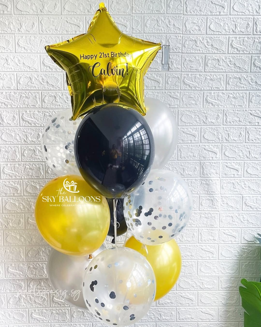 A balloon bouquet featuring a large gold star-shaped foil balloon with 'Happy 21st Birthday Calvin' printed on it, surrounded by black, gold, white, and clear balloons with confetti details, set against a white textured brick wall.
