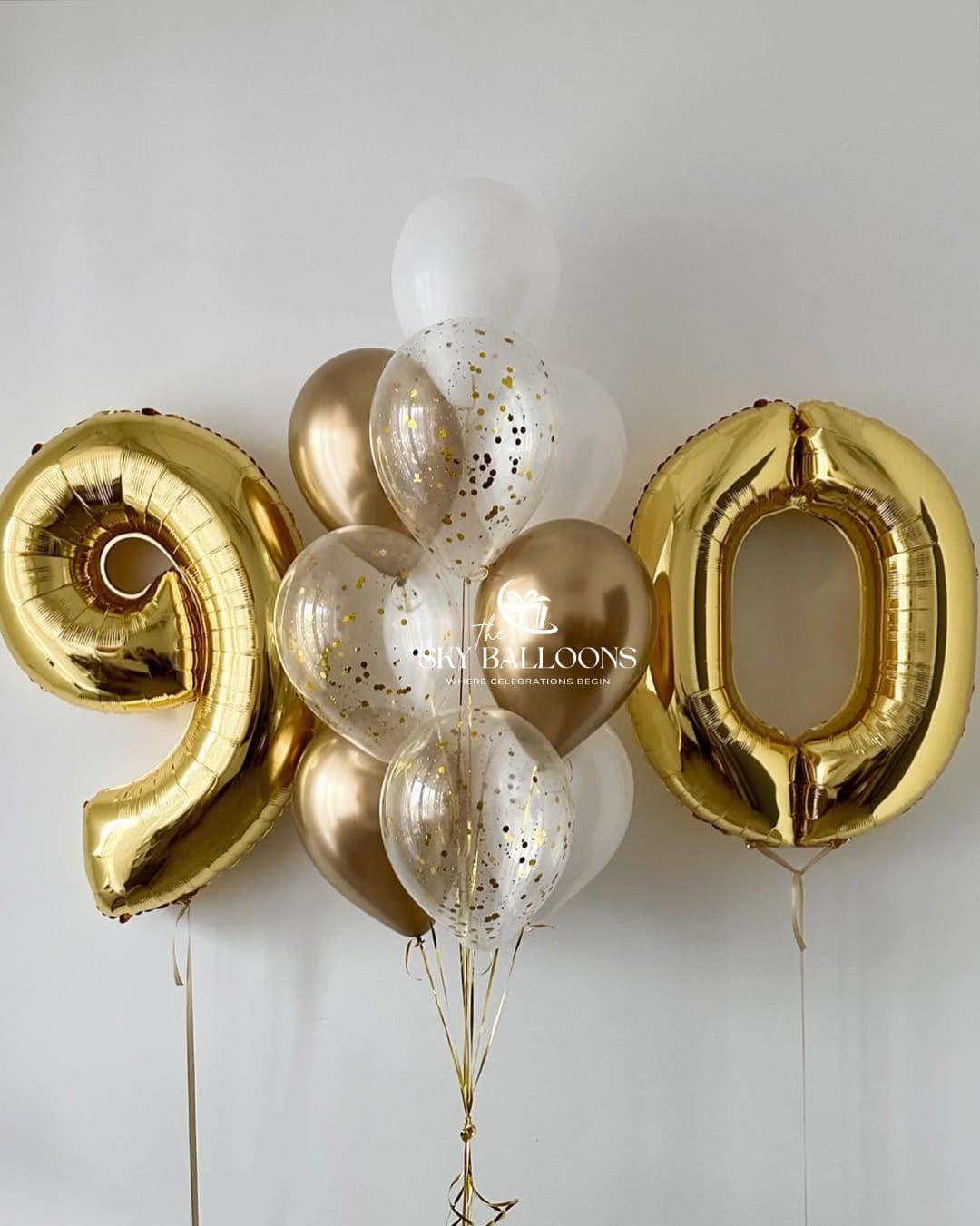 Gold number 90 foil balloons framing a bouquet of white, metallic gold and clear gold confetti balloons with trailing gold ribbons.