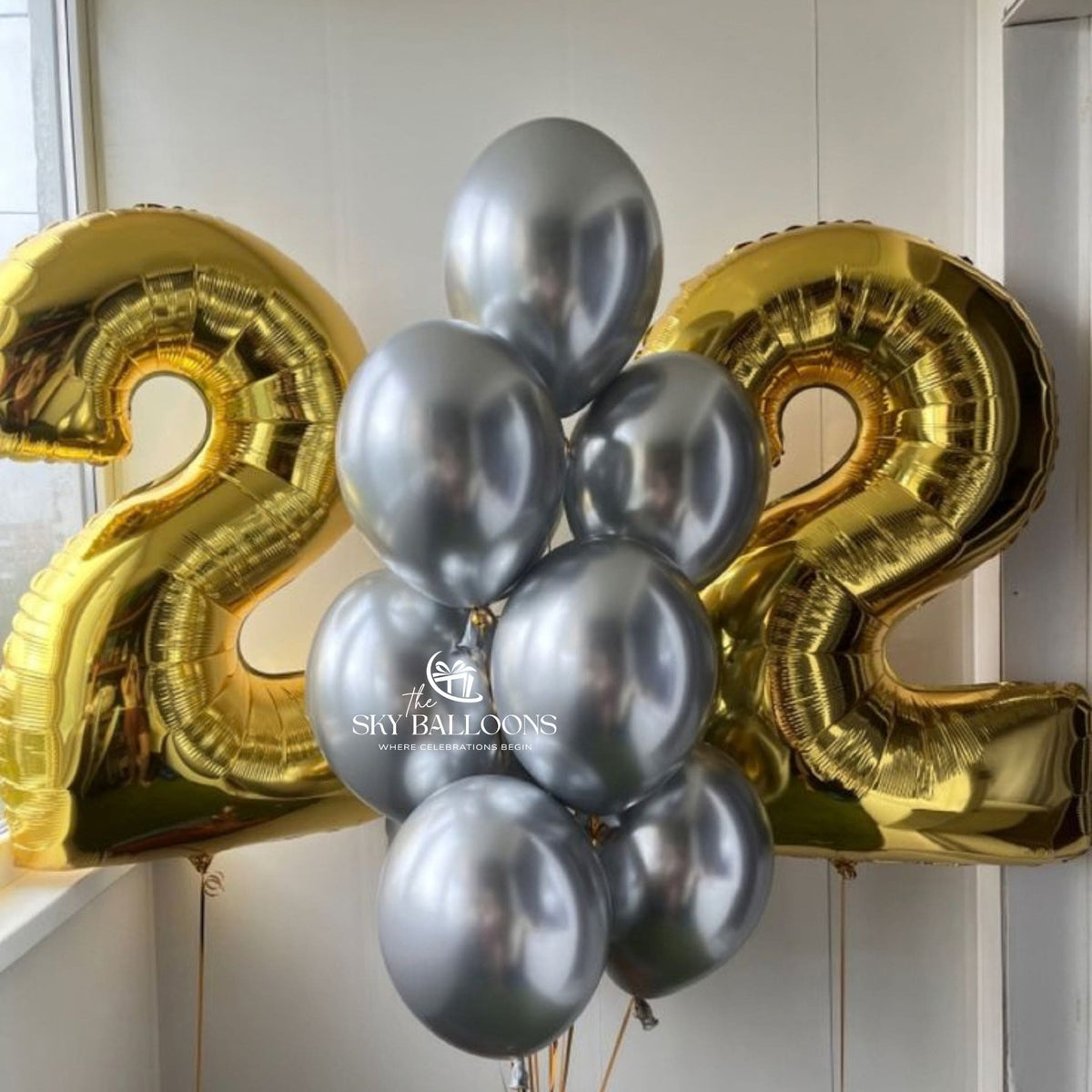 Gold number 22 foil balloons framing a bouquet of metallic silver latex balloons with trailing ribbons against a white wall.