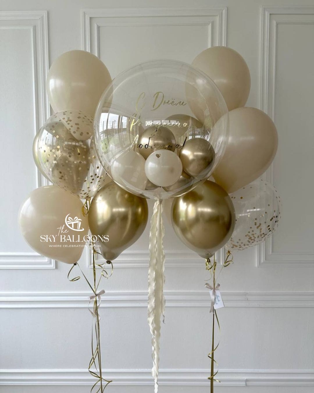 Personalised clear bubble balloon filled with small white and gold balloons, flanked by cream, metallic gold and clear gold confetti balloon bouquets.