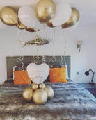 Decorative setup with gold and white balloons and a heart-shaped pillow on a bed.