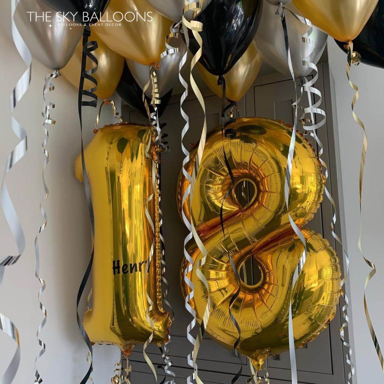 Gold number 18 balloons with silver ribbons against a white wall.