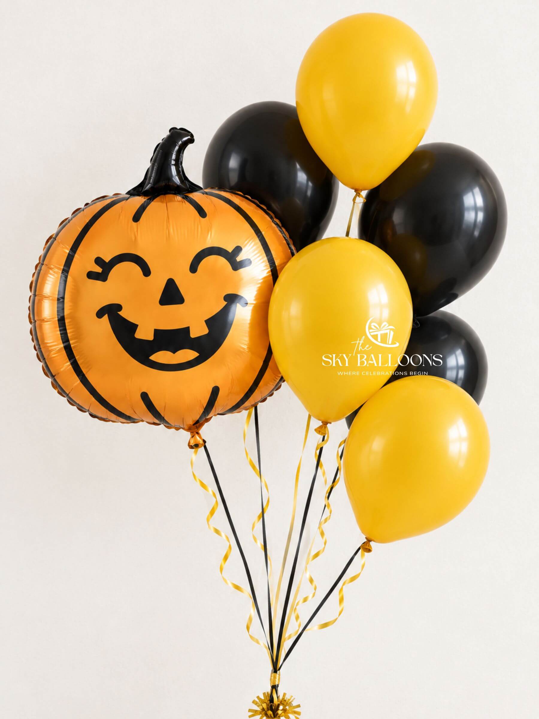 A cluster of Halloween-themed balloons including a large orange jack-o'-lantern foil balloon with a black face, several matte black latex balloons, and four yellow latex balloons, tied together with black and gold ribbons against a plain light background.