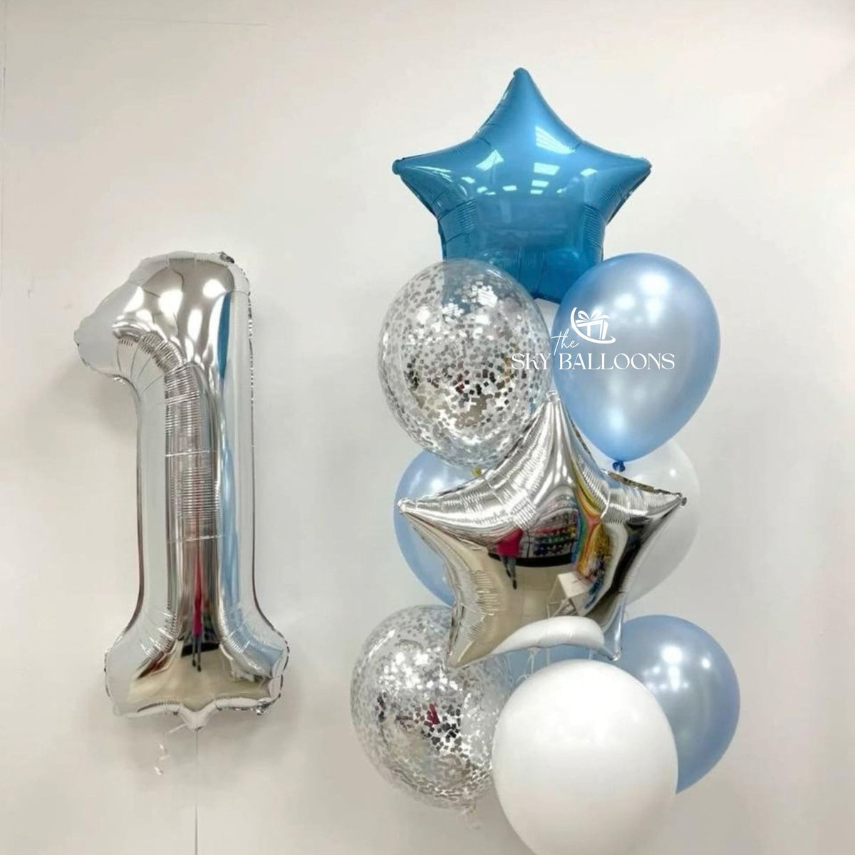 Silver number 1 foil balloon beside a bouquet of blue and silver stars, light blue and white latex balloons, and clear silver confetti balloons.