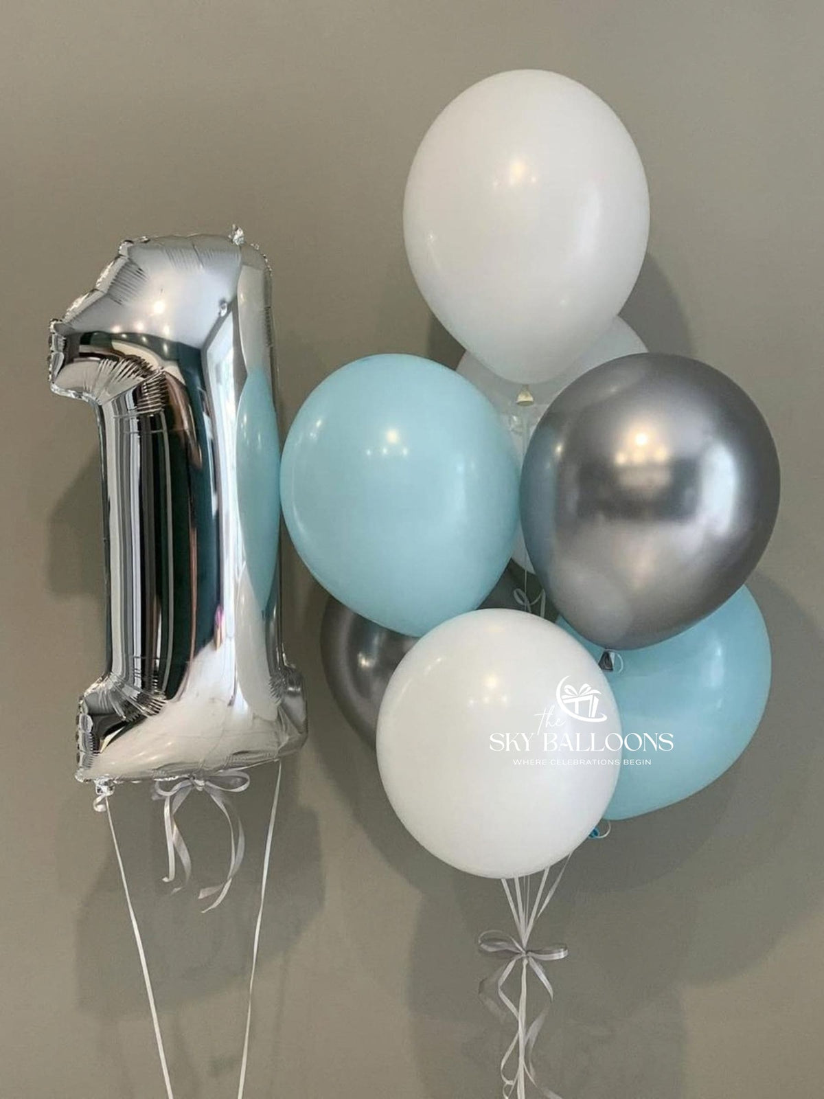 Silver number 1 foil balloon beside a bouquet of light blue, white and metallic silver latex balloons tied with white ribbons.