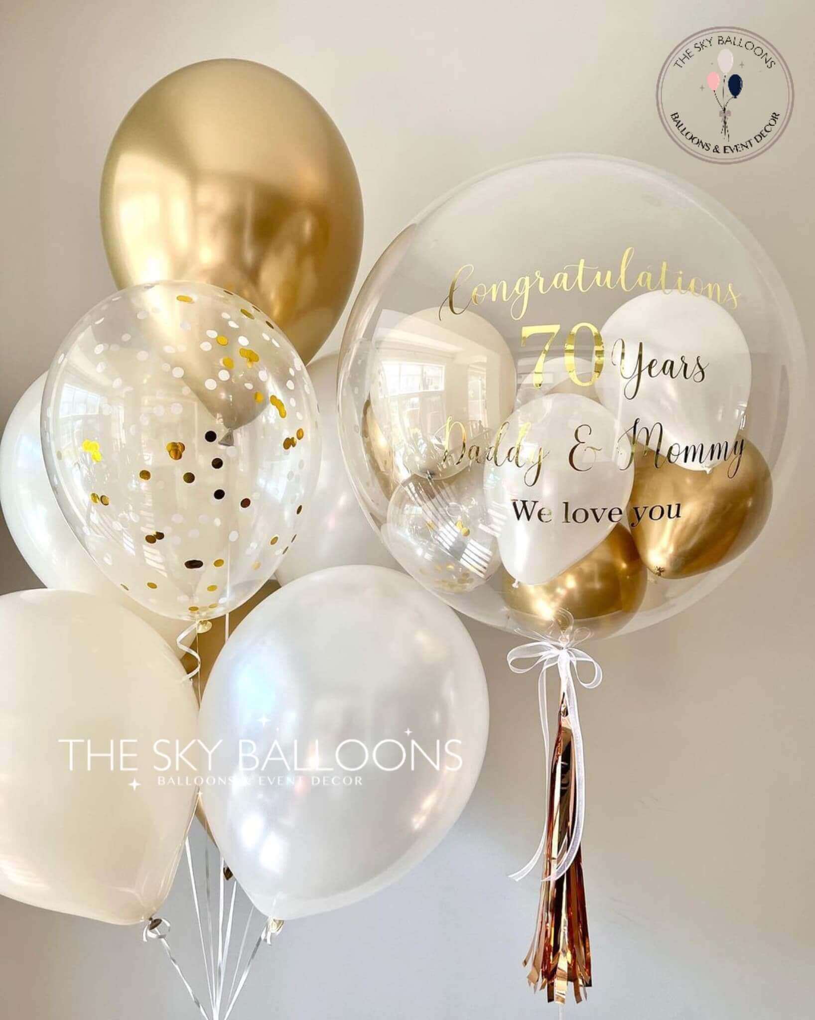 A bouquet of latex balloons filled with helium gas, featuring white and gold colors with printed text that reads 'Happy 10th Anniversary'. There are also smaller balloons and tassels visible within the larger balloons.