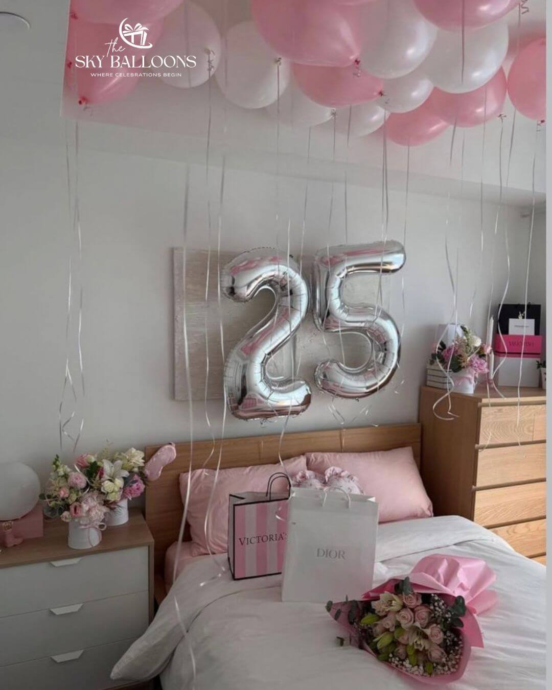25th birthday bedroom decoration with silver number balloons, pink and white ceiling balloons with hanging ribbons, flowers and gift bags on the bed.