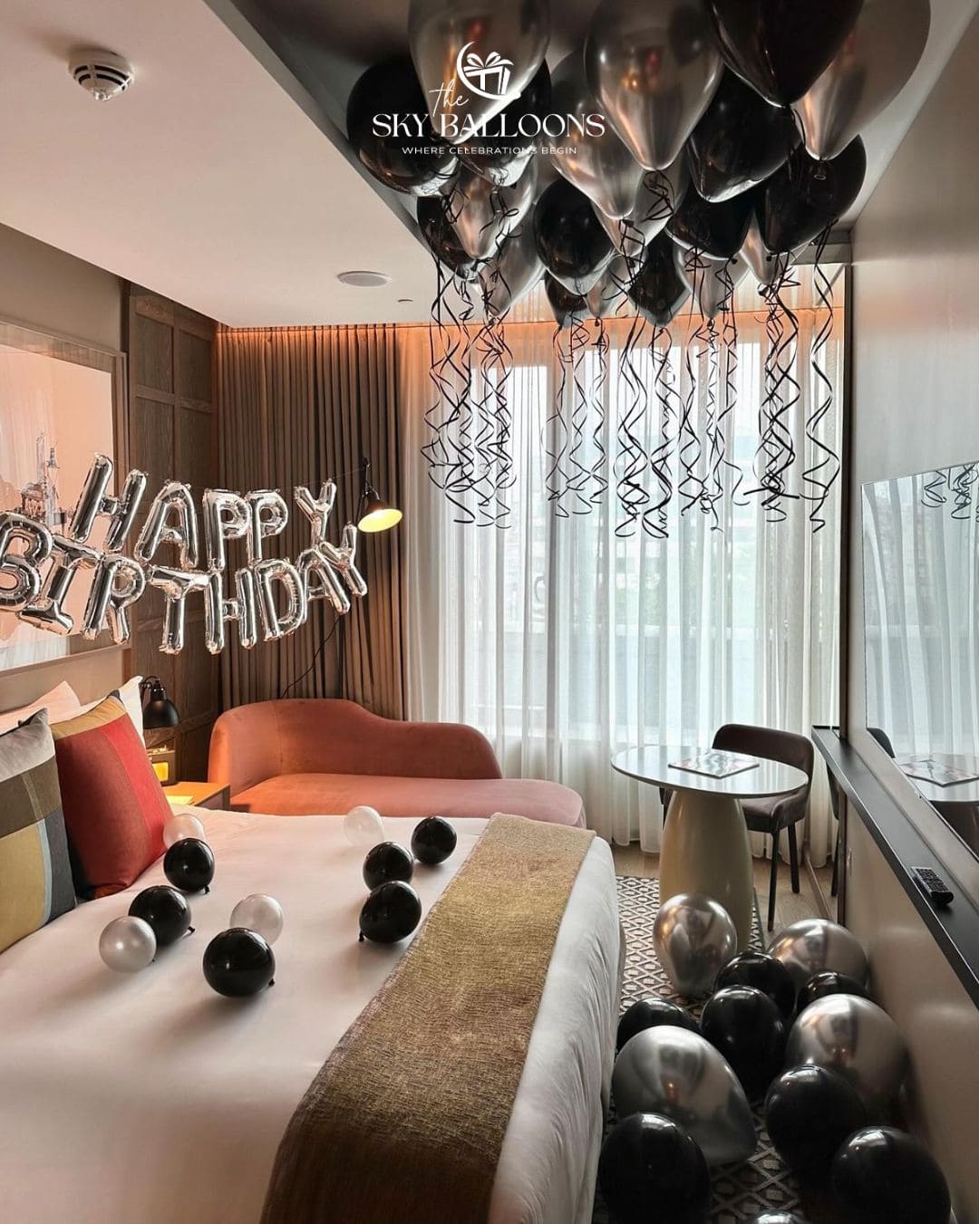 A decorated bedroom with black and silver balloons, black and silver streamers, a large silver 'HAPPY BIRTHDAY' balloon sign, and a bed with black and silver balloons on the bedding. The room has a modern design with a pinkish-orange curved armchair, white curtains, and a window.