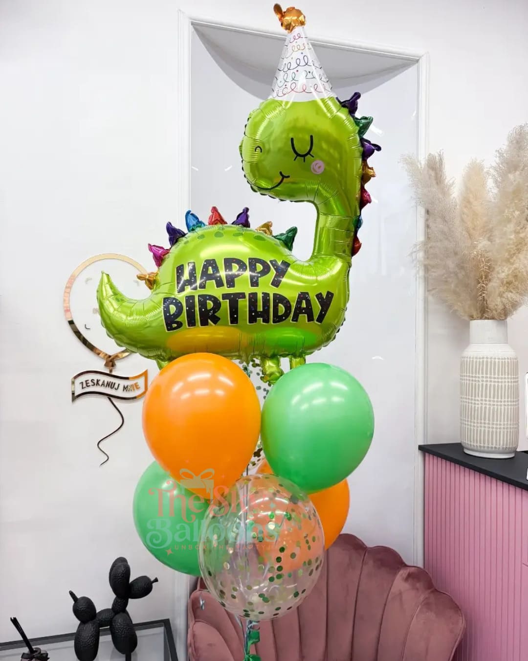 Green dinosaur-shaped balloon with 'Happy Birthday' text, surrounded by other balloons, against a white wall.