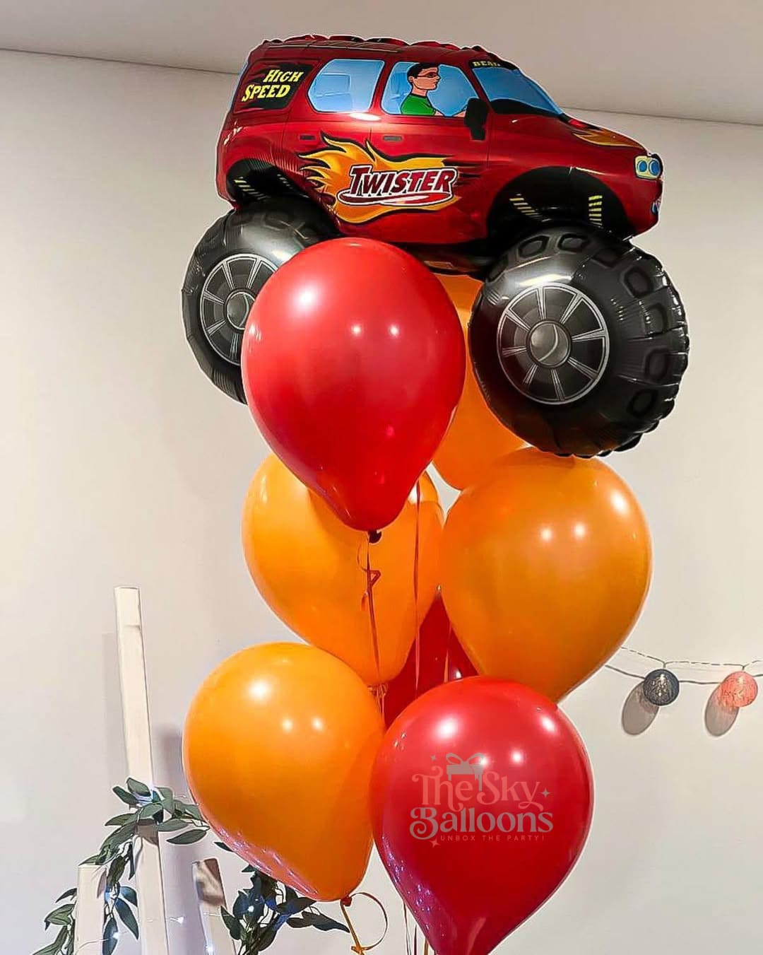 Toy car with large wheels surrounded by red and orange balloons against a white wall.