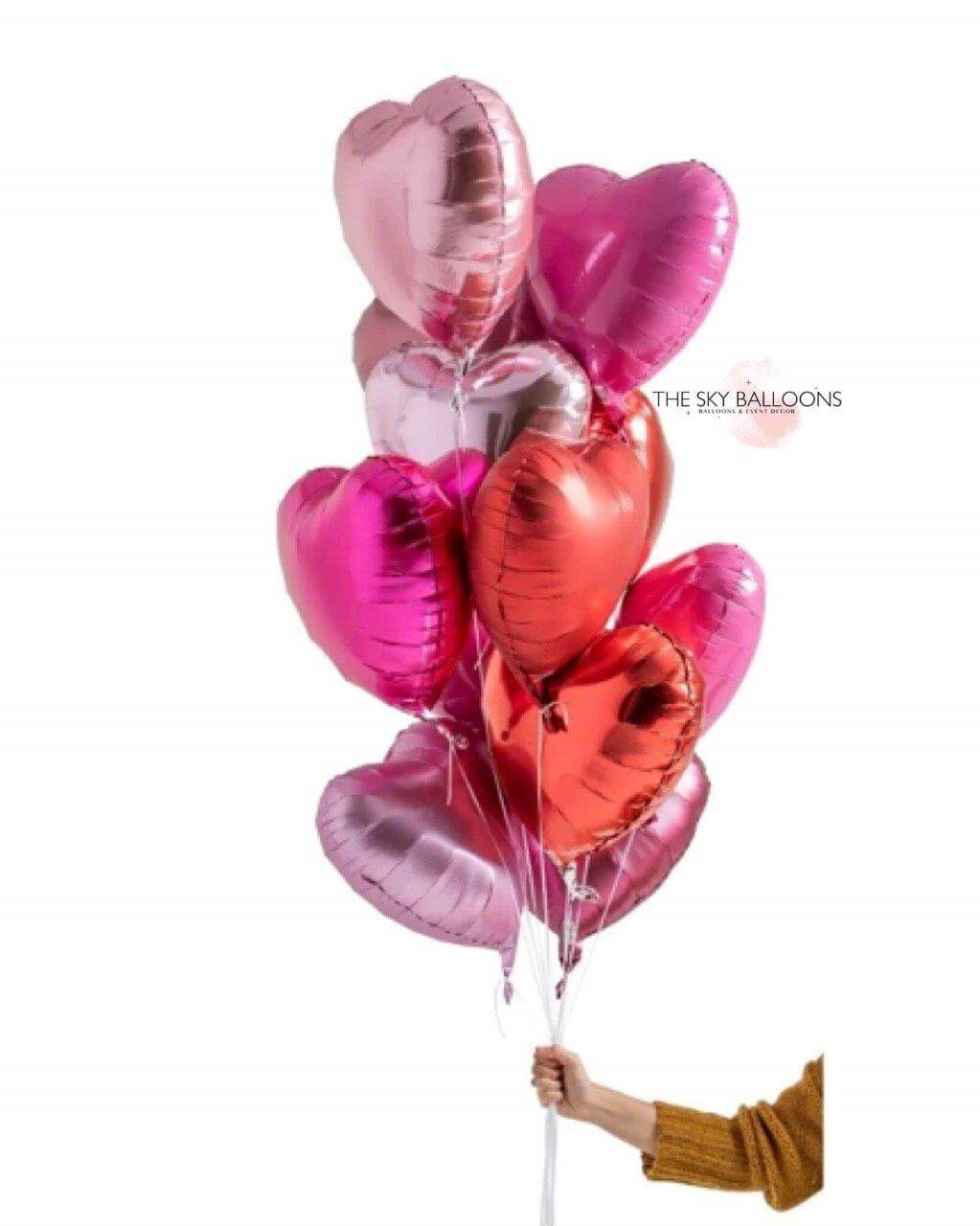A bouquet of heart-shaped balloons in various shades of pink, silver, and gold, held by a hand against a white background.
