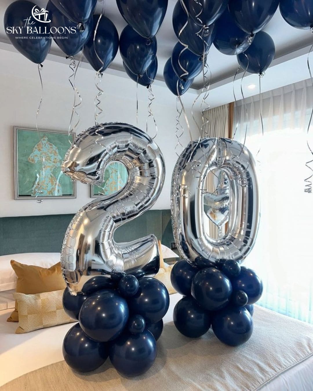 Two large metallic silver number 2 and 0 foil balloons, each mounted on a cluster of dark navy blue balloons, displayed on a bed in a modern bedroom. More dark navy balloons are floating around the room.