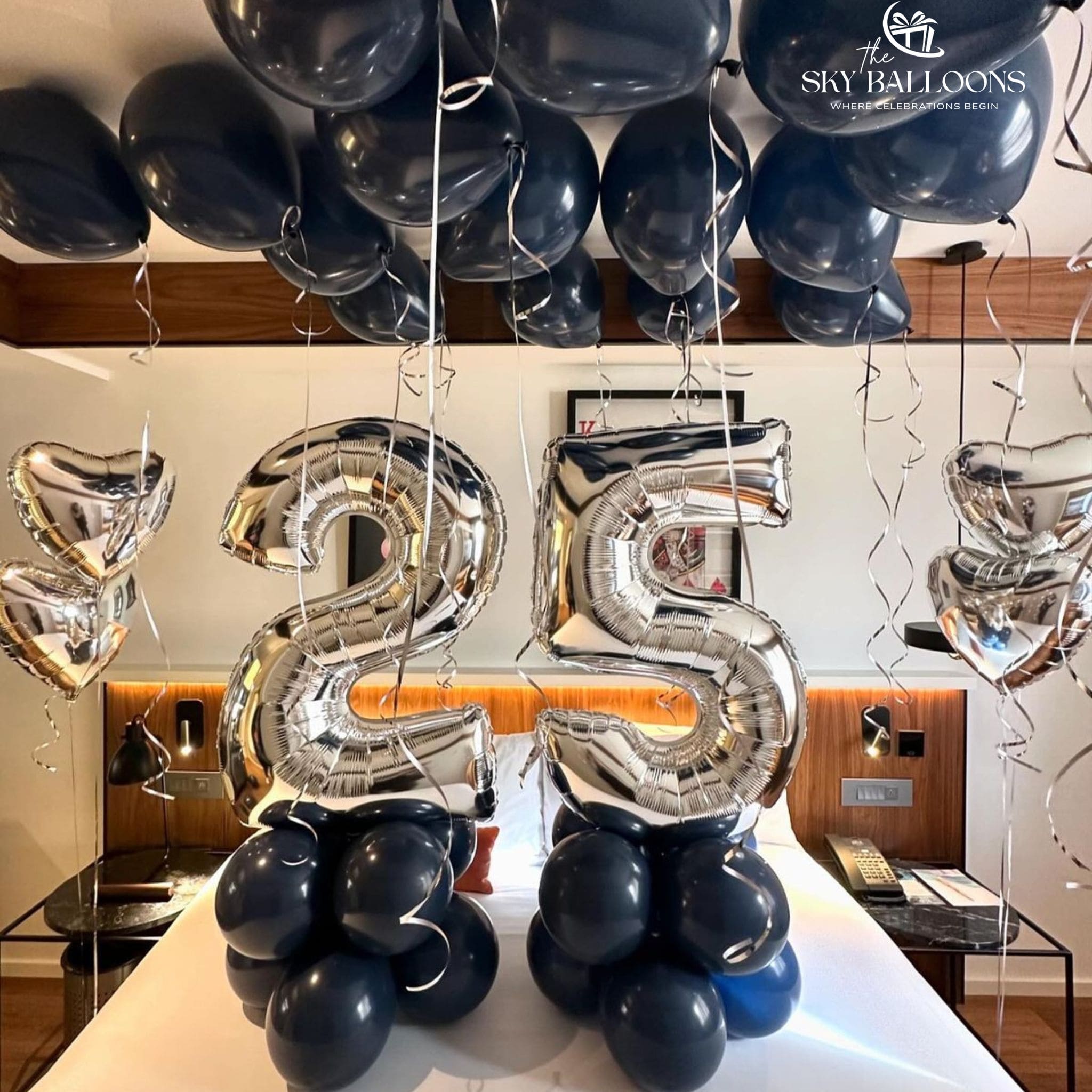 Silver foil number balloons spelling '25' displayed on a bed in a hotel room, surrounded by navy blue latex balloons and hanging balloons from the ceiling. The 'The Sky Balloons' logo is visible in the top right corner.