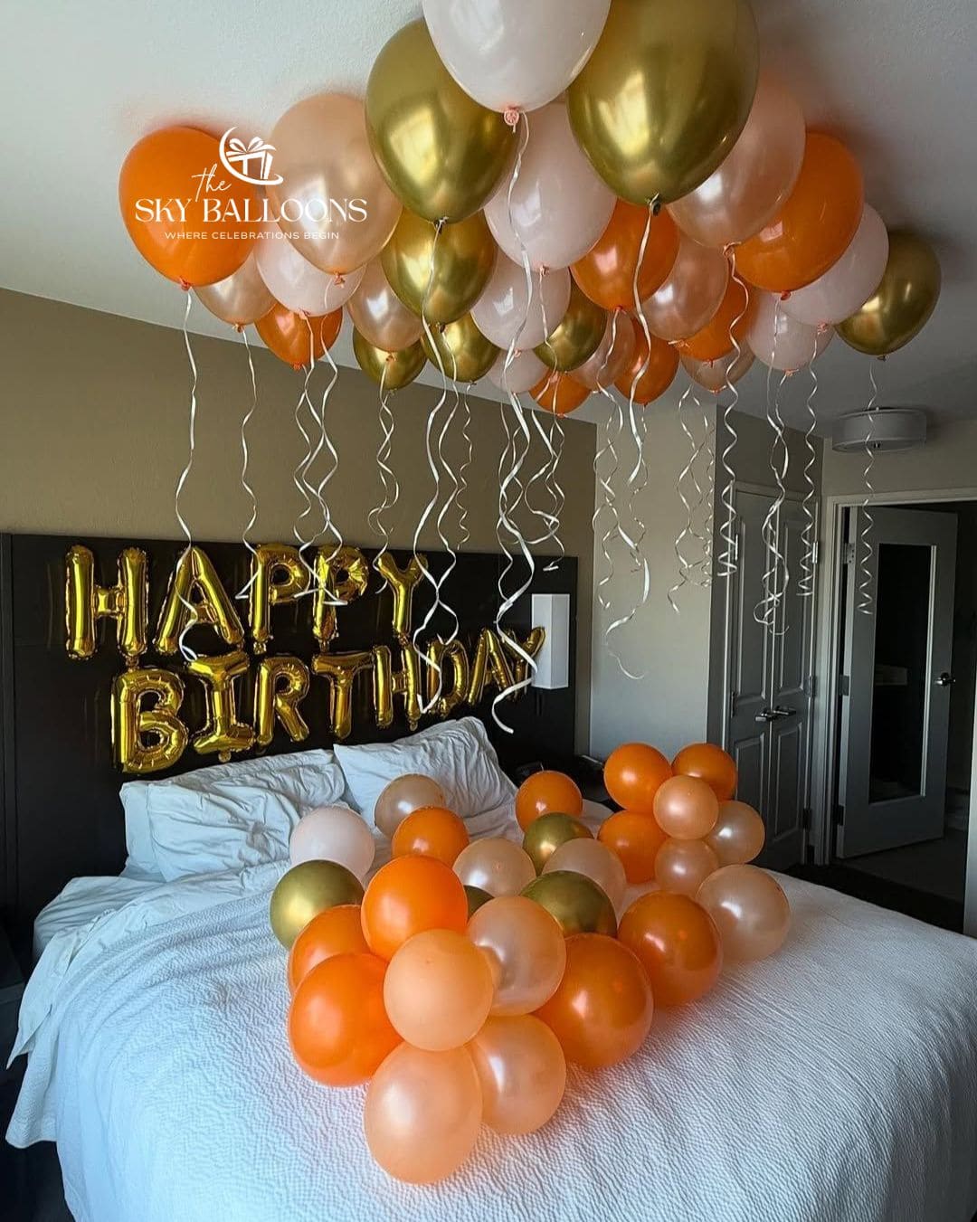 A bedroom decorated for a birthday with a cluster of metallic gold, orange, and blush pink balloons floating above the bed, with a matching balloon garland arranged on the white bed. Gold foil 'HAPPY BIRTHDAY' letters are displayed on the headboard, and a watermark for 'The Sky Balloons' is visible in the upper left.
