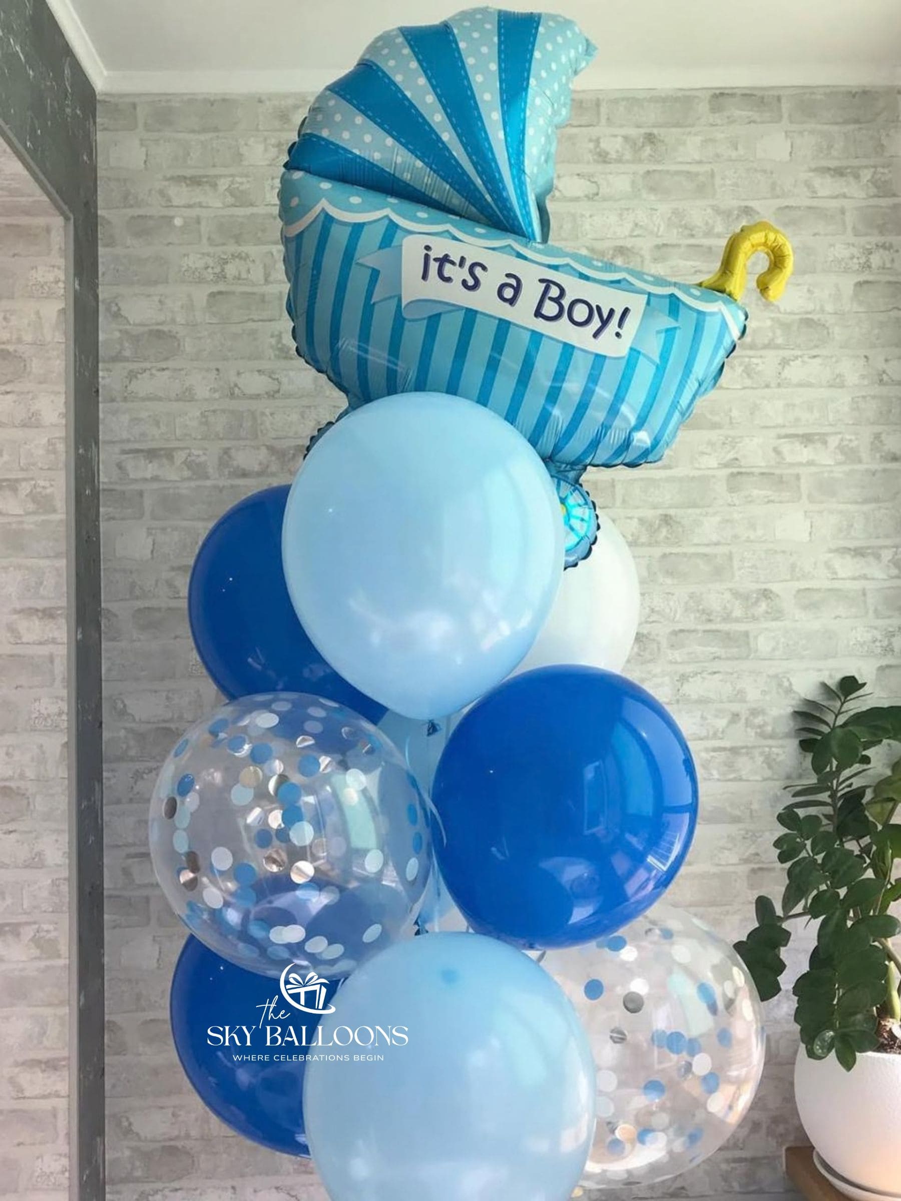 A baby shower decoration balloon cluster featuring a blue and white striped foil balloon shaped like a baby stroller with the text 'it's a Boy!' and a yellow rattle, surrounded by blue, light blue, white, and clear balloons with confetti, set against a gray brick wall.