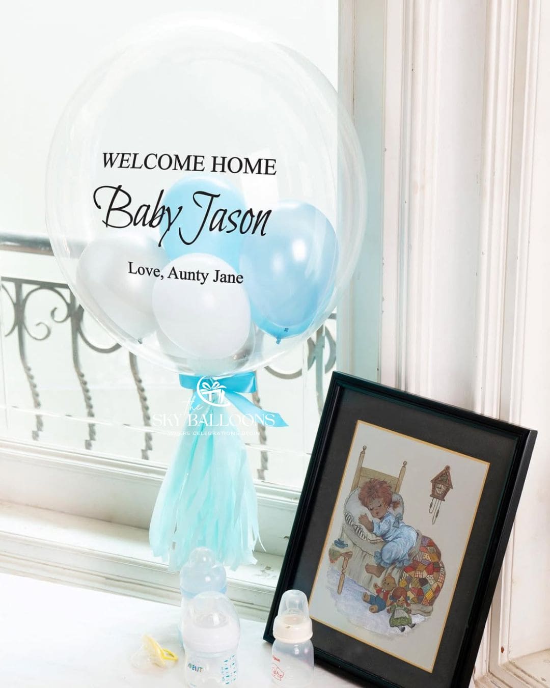 Personalised clear bubble balloon reading “Welcome Home Baby Jason, Love, Aunty Jane,” filled with blue and white balloons and finished with a blue bow and tassels.