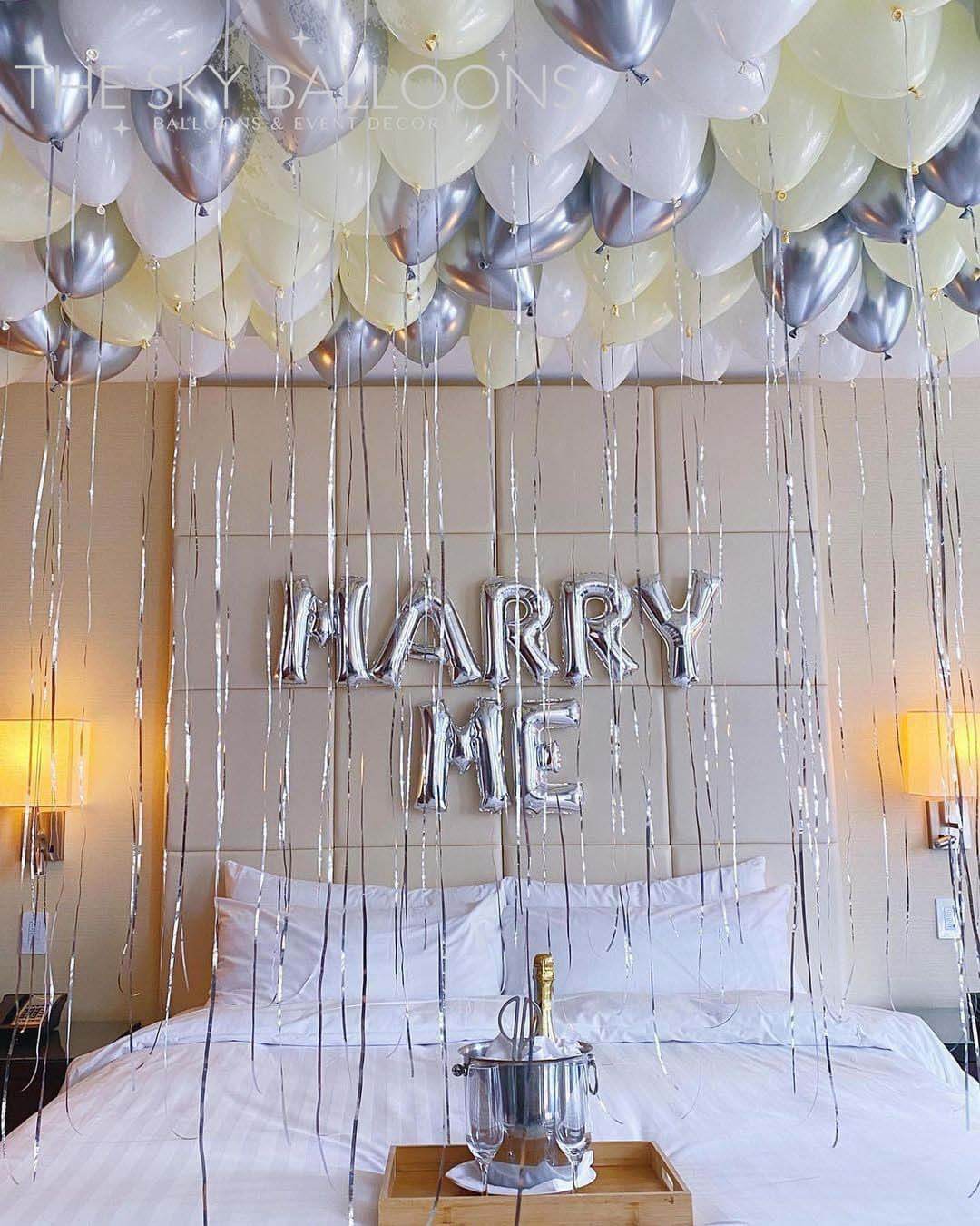 A bedroom setup with silver, chrome, ivory, and white balloons forming a 'MARRY ME' banner above the bed.