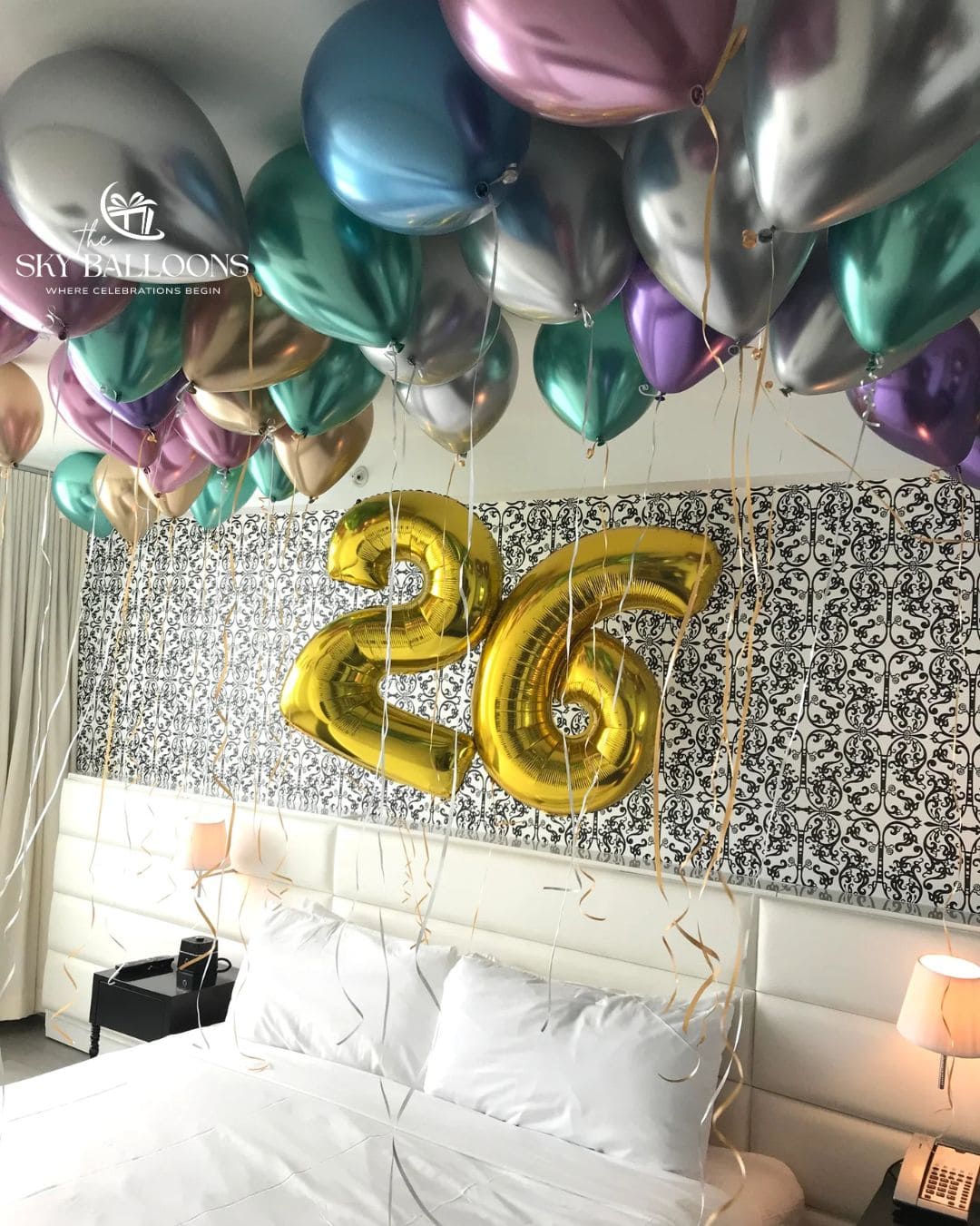 A bedroom scene decorated for a birthday with a bed, patterned wall, bedside lamps, and many metallic balloons in silver, blue, purple, and green, plus large gold foil numbers reading 26.
