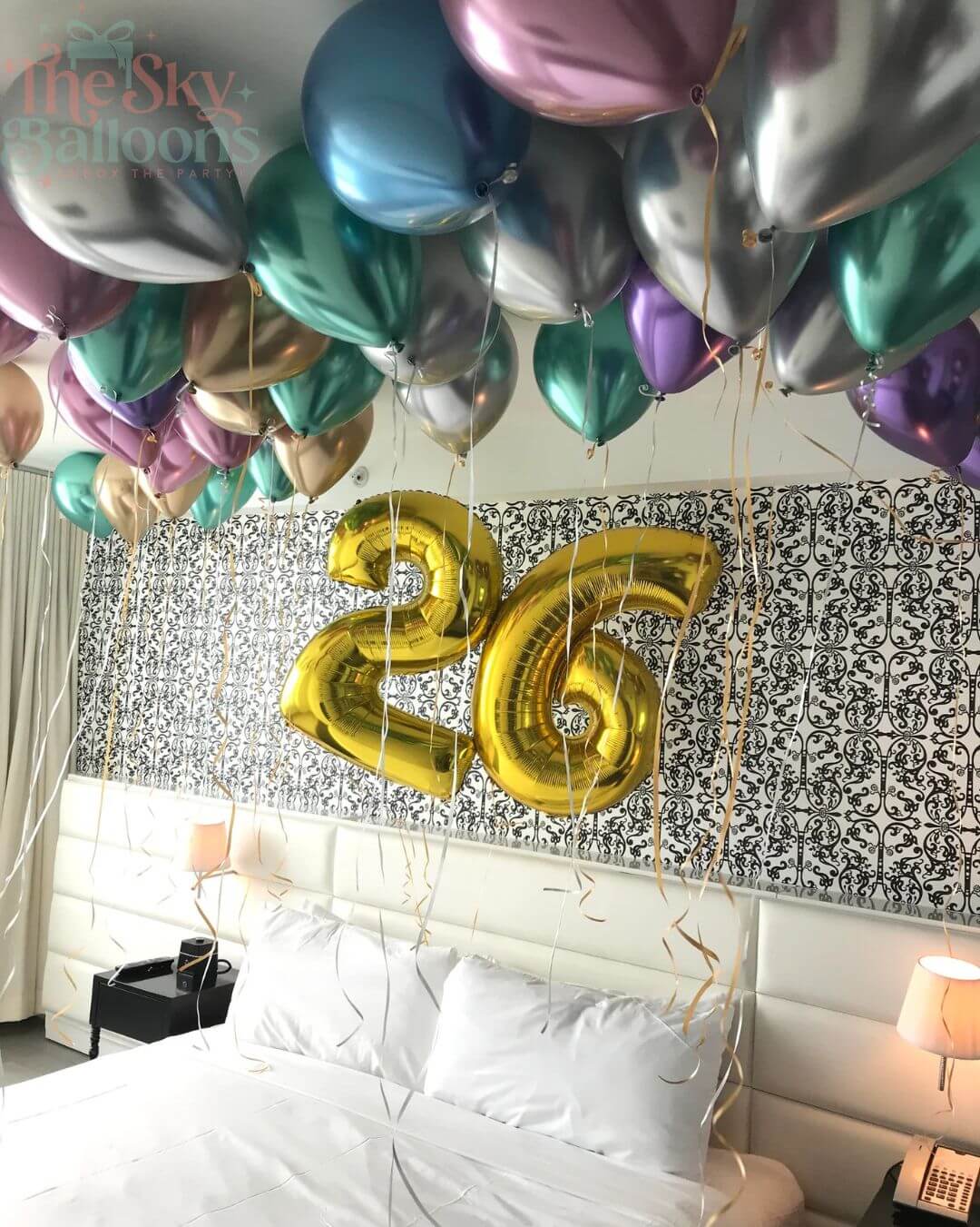 Gold number '26' balloons in front of a bed with decorative wall and 'The Sky Balloons' branding.