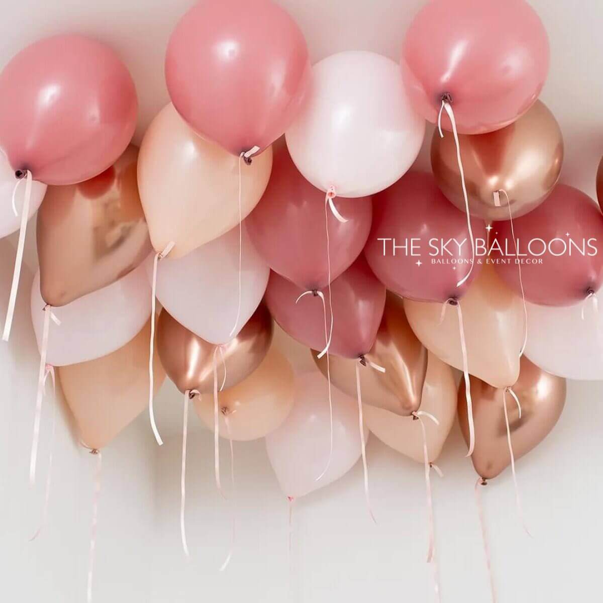 A cluster of inflated balloons in shades of pink, rose gold, and white, attached to strings, with the brand name 'The Sky Balloons' visible.