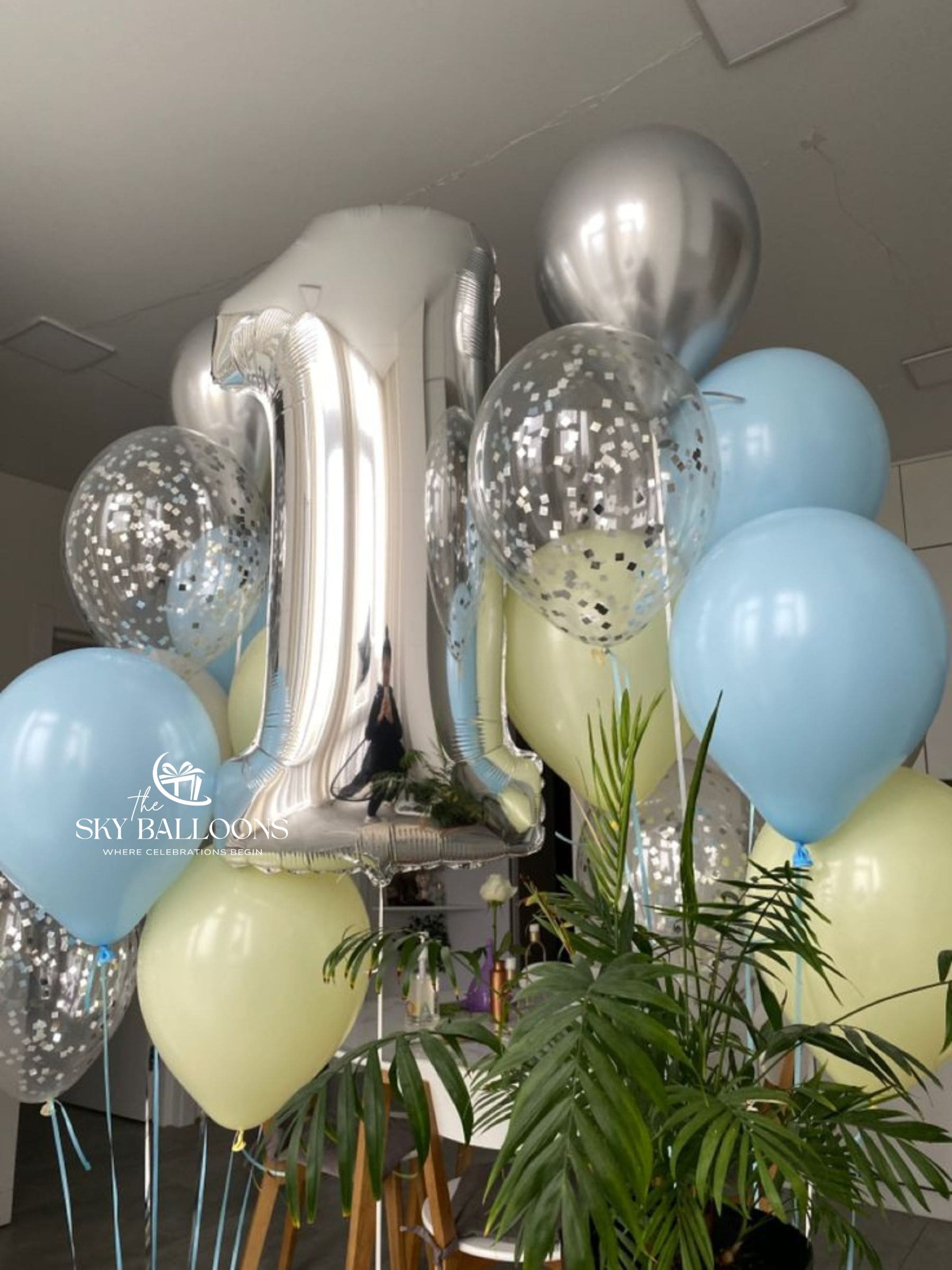 Two pastel yellow, pastel blue, chrome silver and silver-confetti balloon bouquets with a large silver number 1 foil balloon for a birthday celebration.