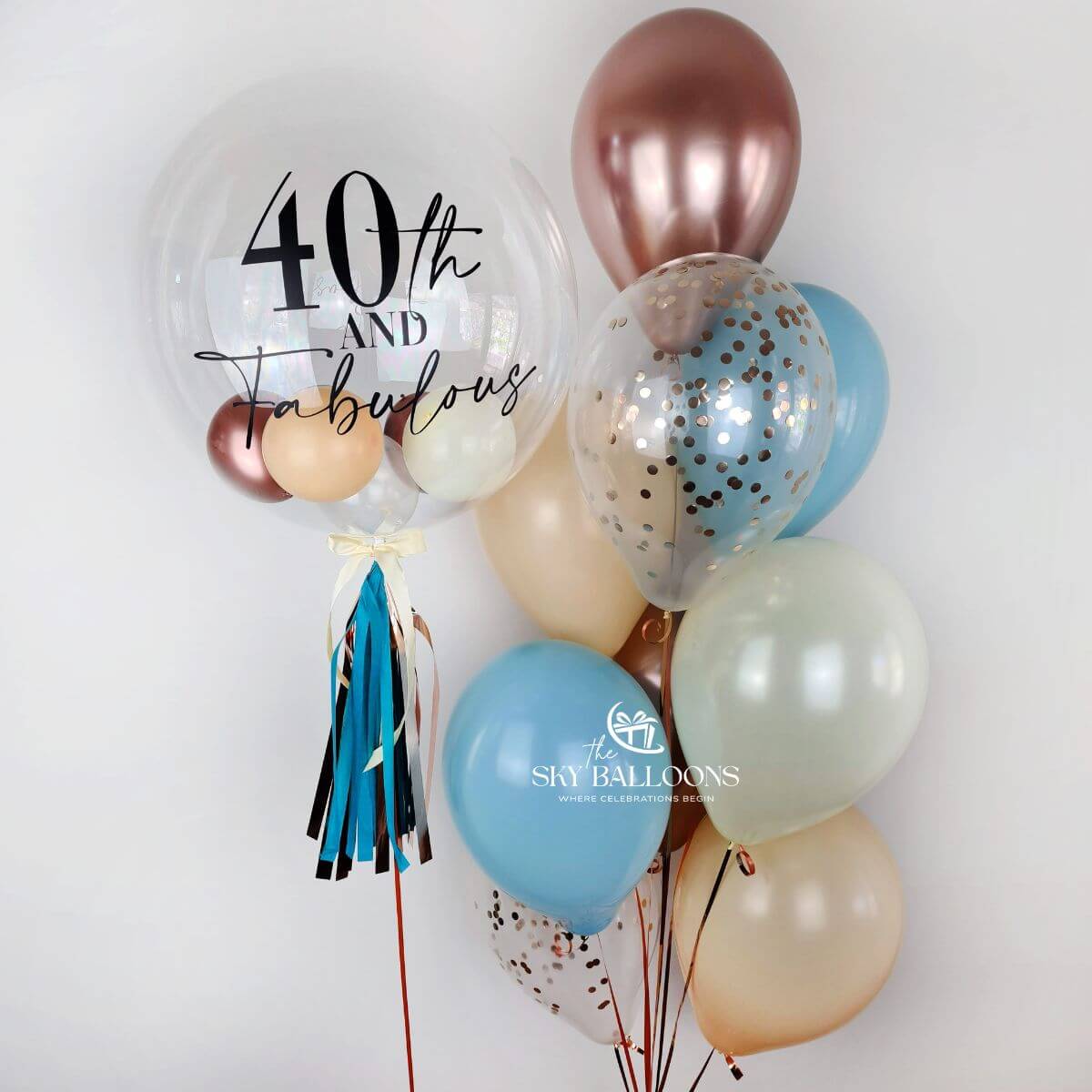 Personalised clear birthday bubble balloon with decorative tassels beside blue, rose-gold, cream and clear confetti balloons.