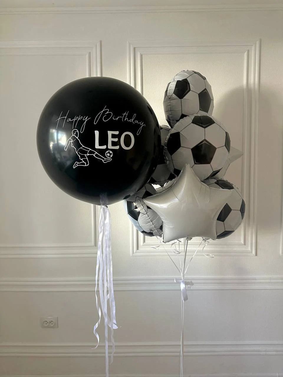 Black balloon with 'Happy Birthday Leo' text, soccer ball balloons, and star balloon against a white paneled wall.