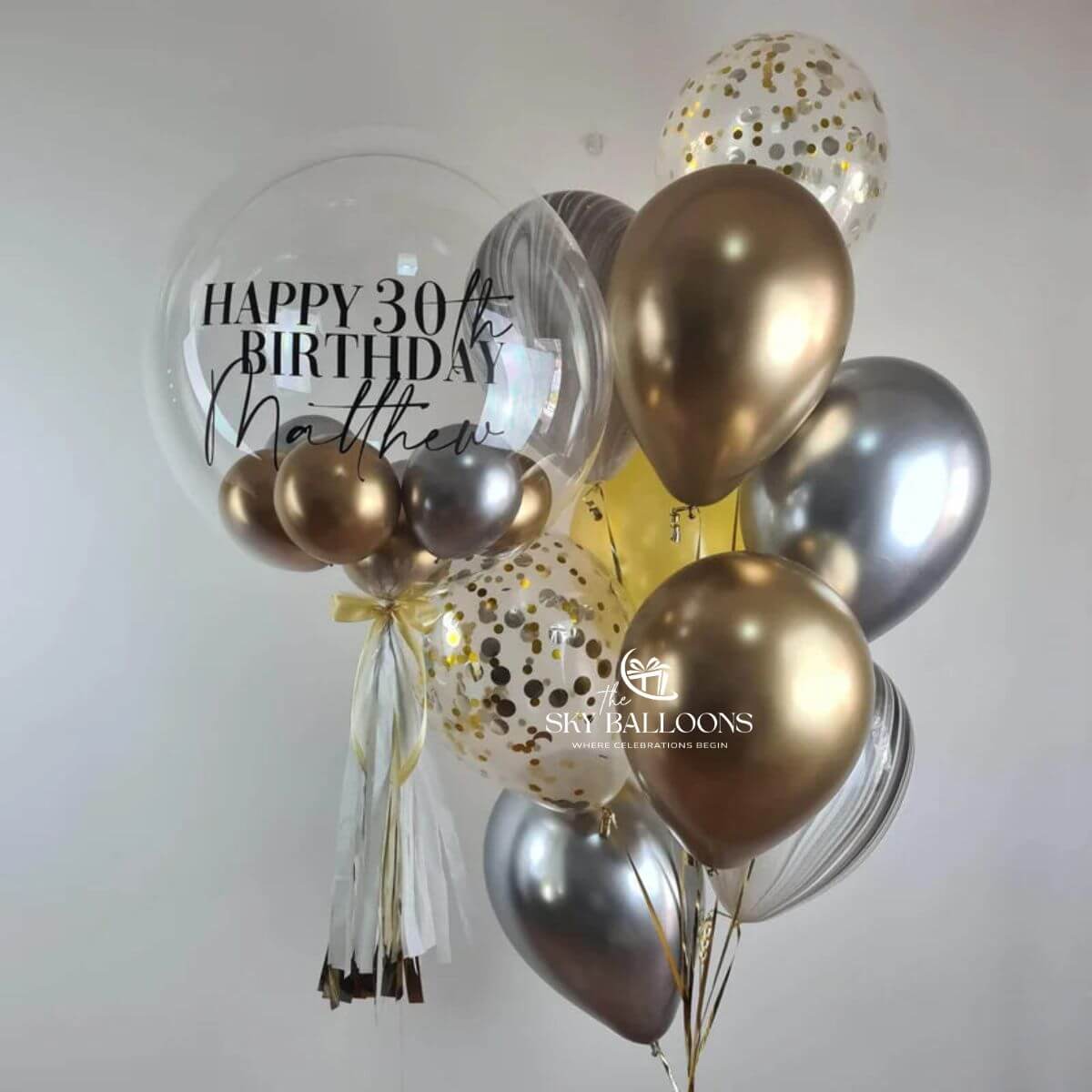 Personalised clear birthday bubble balloon with gold and silver mini balloons and tassels beside metallic, marble-effect and gold-confetti balloons.