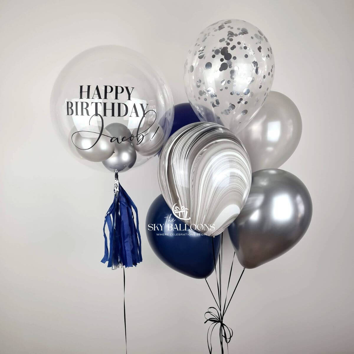 Personalised clear birthday bubble balloon with navy tassels beside navy, silver, marble-effect and clear silver-confetti balloons.