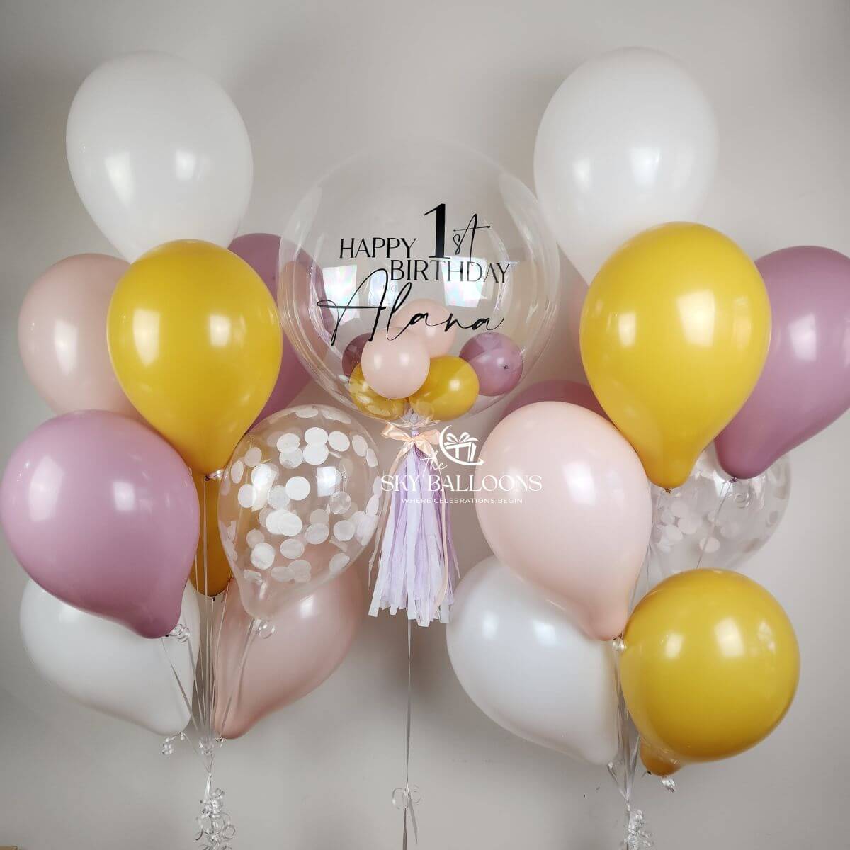 Two pink, lilac, mustard-yellow, white and confetti balloon bouquets beside a personalised clear birthday bubble balloon with tassels.