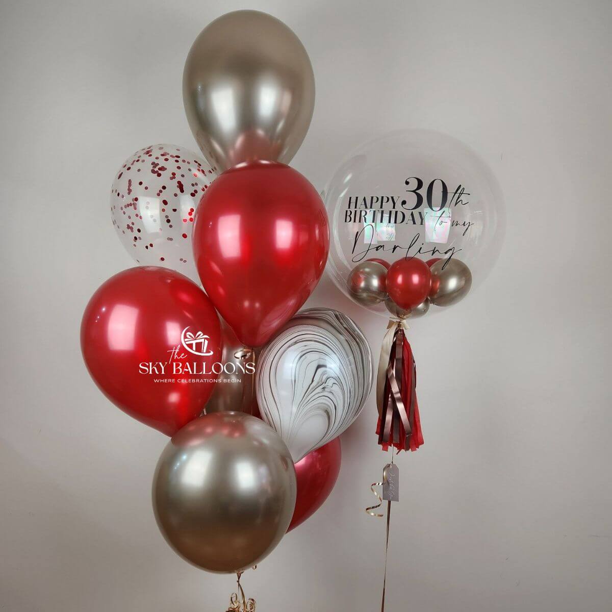 Personalised clear birthday bubble balloon with red and silver mini balloons and tassels beside red, silver, marble-effect and confetti balloons.
