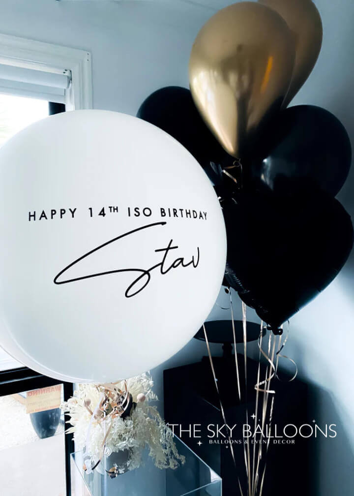 A bouquet of balloons featuring black and gold colored latex balloons with one personalized balloon that has text on it.