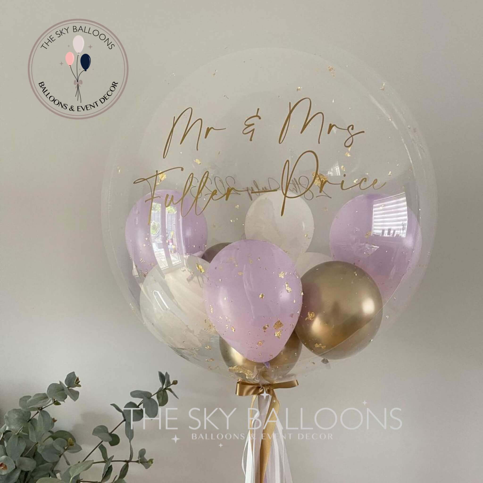 A customized pastel lilac bubble balloon with gold and purple tassels, filled with helium, alongside smaller balloons. There is a golden balloon bouquet with the text 'Mr & Mrs Fullerton Price' written on it.