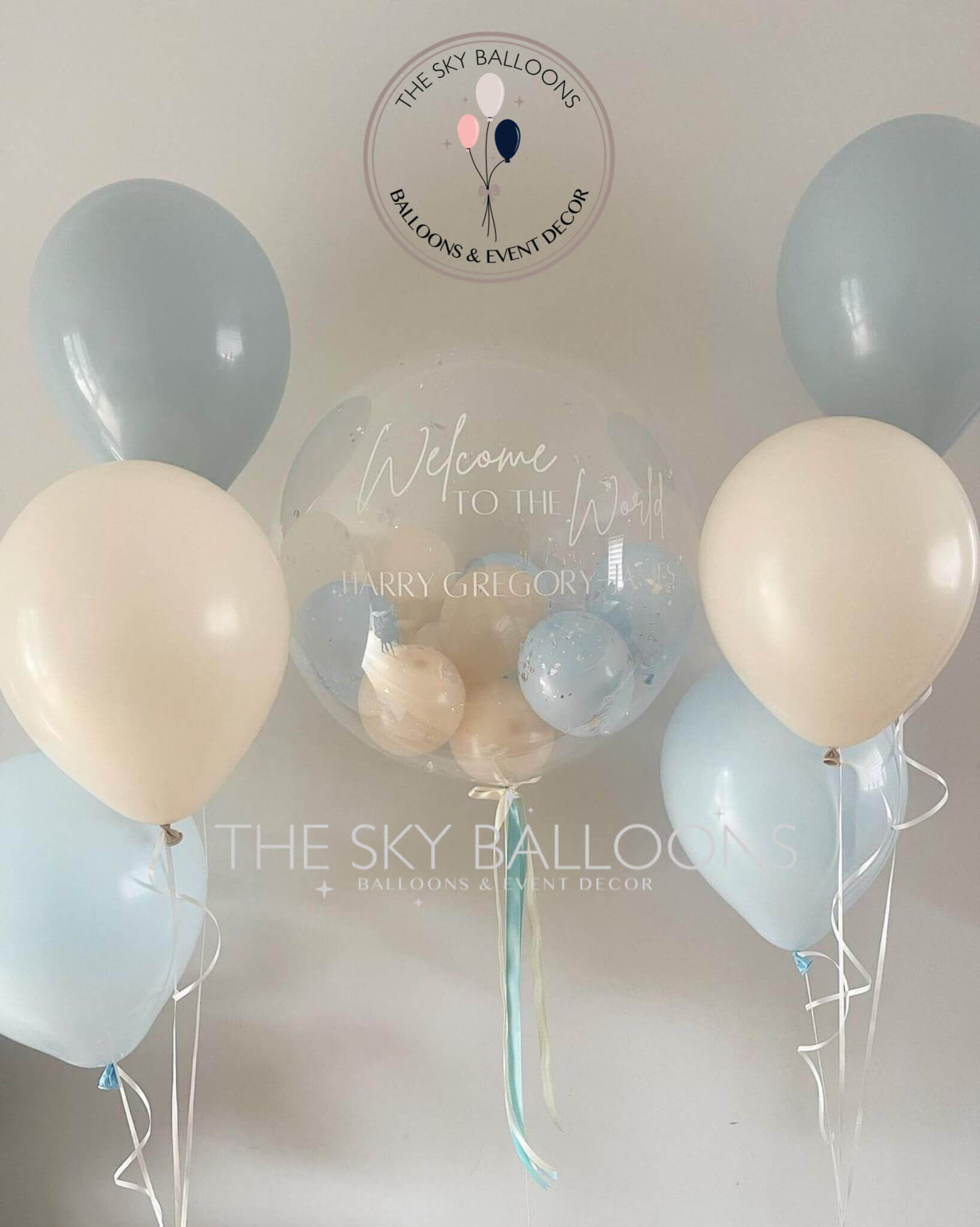 A set of blue latex balloons with a 'Welcome Home' message, including a large balloon with a baby blue and white design, along with several smaller balloons tied to a ribbon.