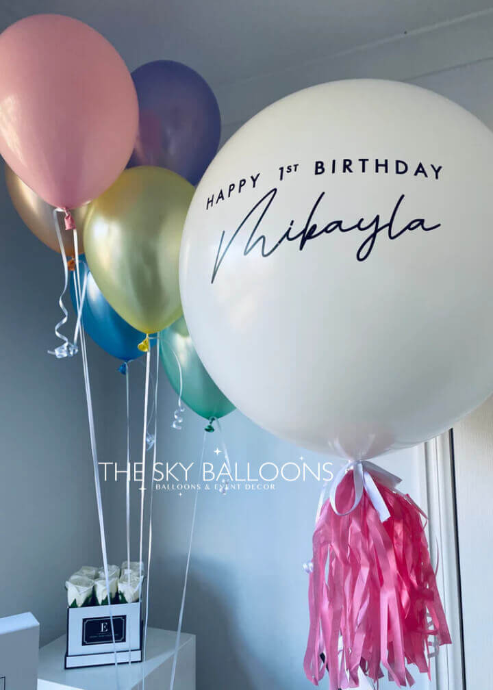 A set of balloons in various colors including pink, gold, blue, and white, with one large personalized balloon reading 'Happy 1st Birthday Mikayla', and attached pink weight tassels.