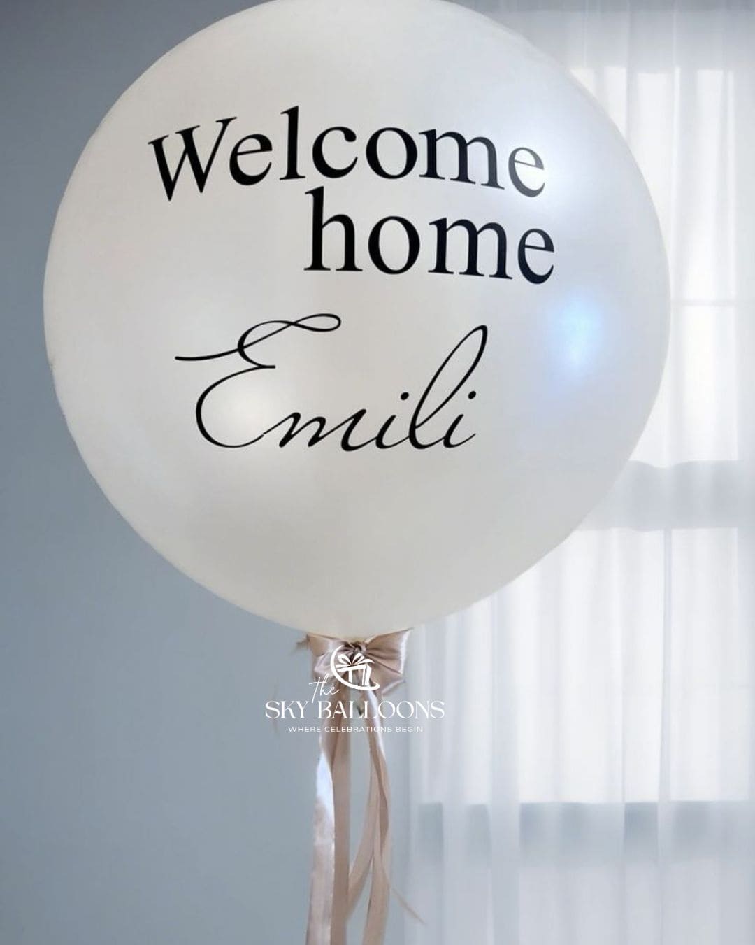 A large white balloon with the words 'Welcome home' and the name 'Emili' printed in black, tied with a pale pink ribbon. A watermark for 'The Sky Balloons' is visible near the bottom.